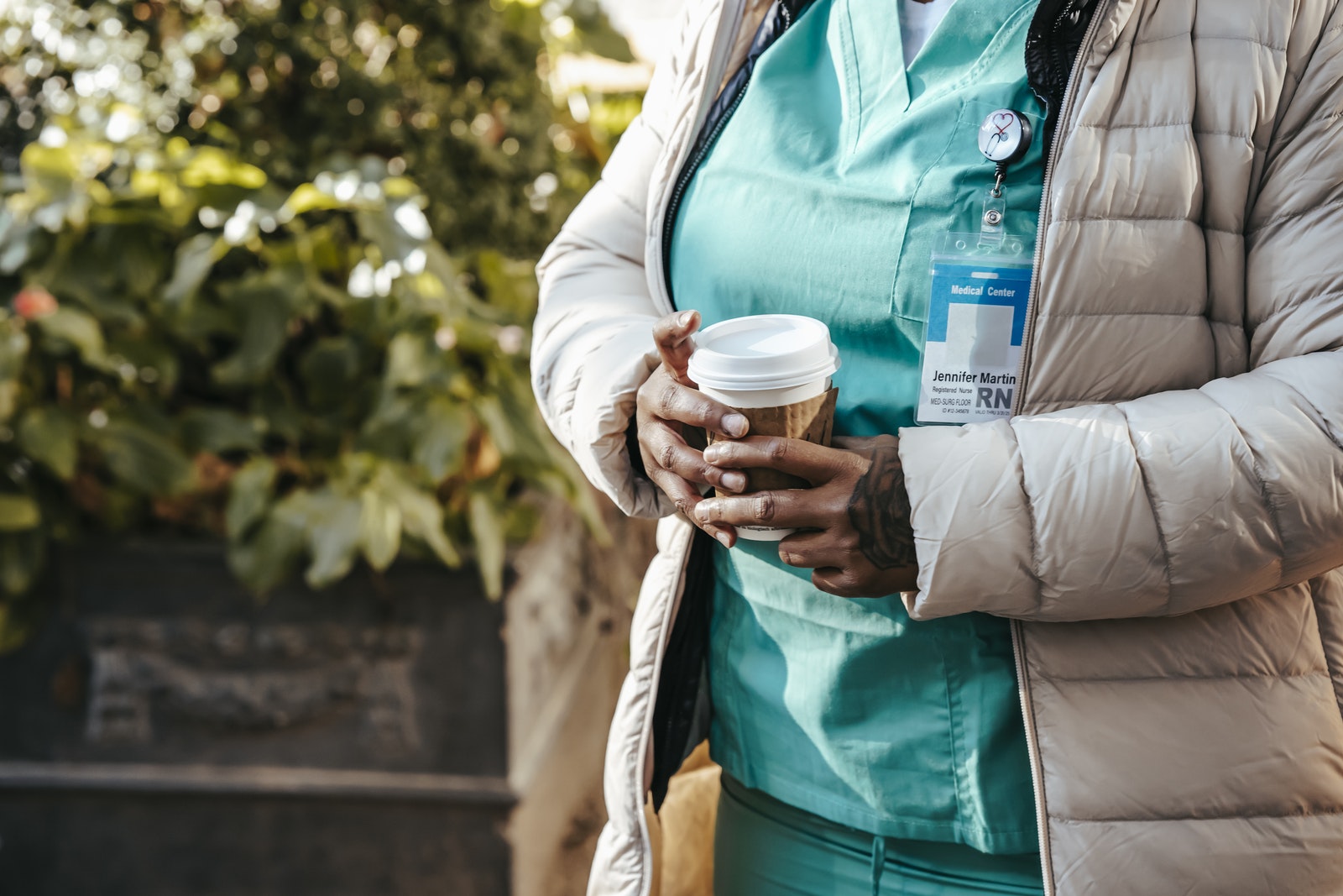 Crop anonymous African American nurse wearing warm clothes standing with coffee to go on street on sunny autumn day