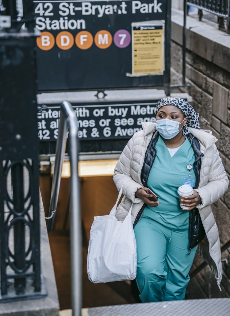 Contemplative African American traveling nurse in outerwear and face mask carrying takeaway coffee and leaving New York metro station while commuting to work