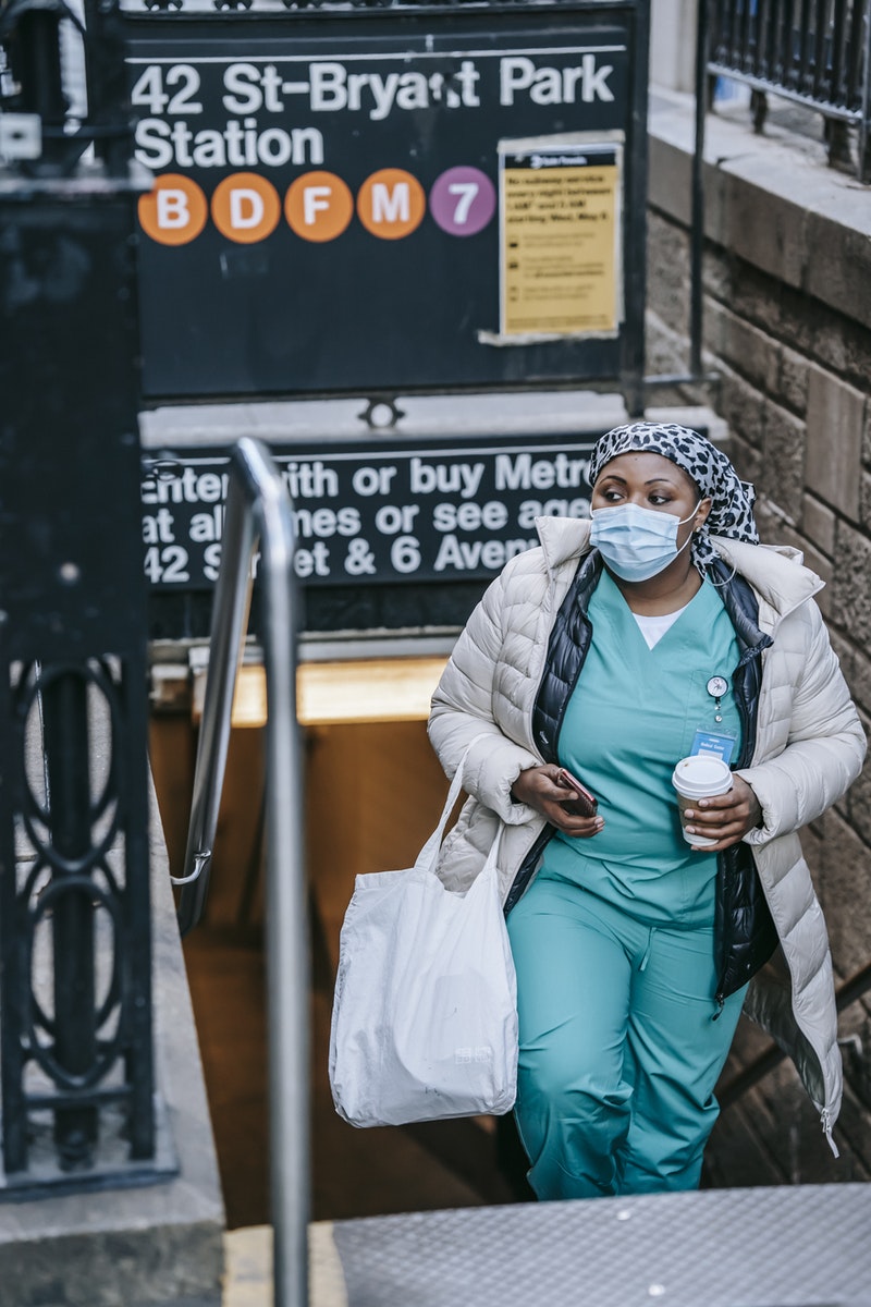 Contemplative African American traveling nurse in outerwear and face mask carrying takeaway coffee and leaving New York metro station while commuting to work