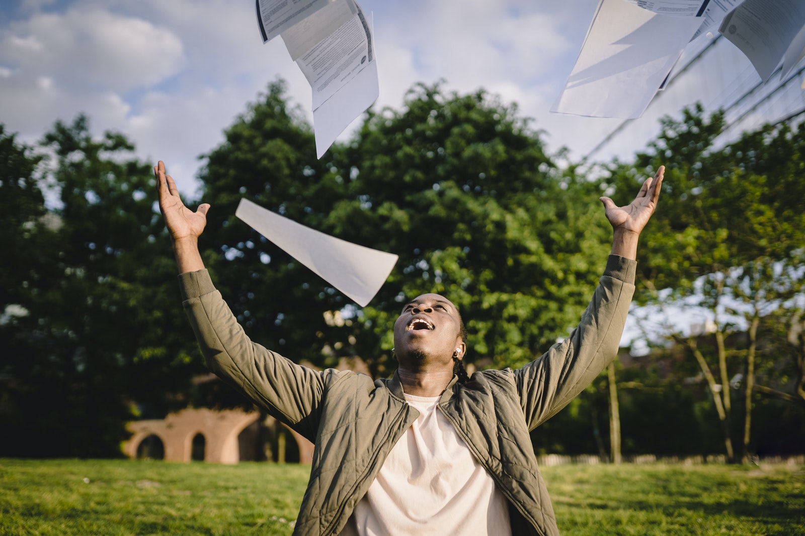 Cheerful young African American male student in casual clothes throwing college papers up in air while having fun in green park after passing anatomy and physiology